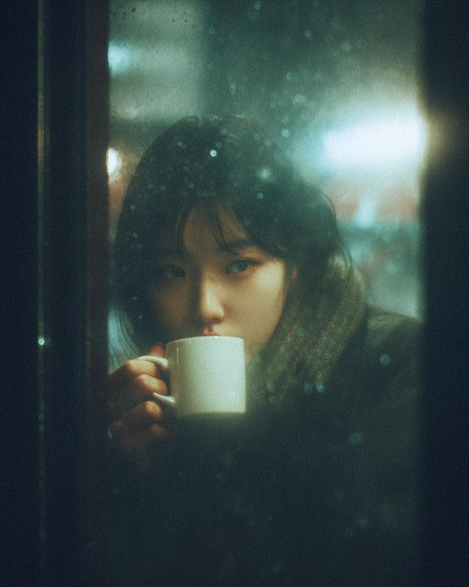 A cinematic, moody photograph of a young woman sitting inside a café at night, seen through a rain-…